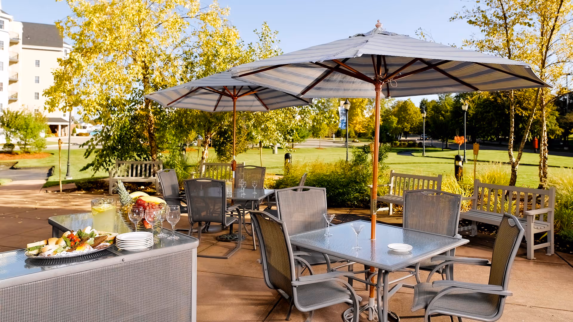 Outdoor patio area with glass tables and chairs under large umbrellas, surrounded by benches and greenery, with a tray of food and drinks on a nearby counter, set in a sunny garden space.