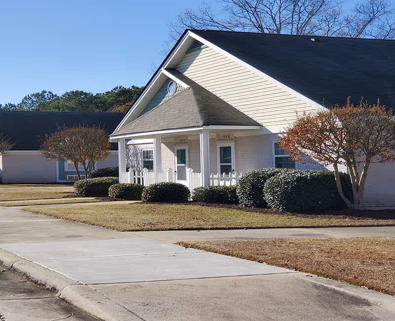 Exterior view of a single-story residential building with a sloped roof, white siding, and a small porch with two doors. The building is surrounded by trimmed bushes and small trees, with a concrete sidewalk and driveway in the foreground under a clear blue sky.