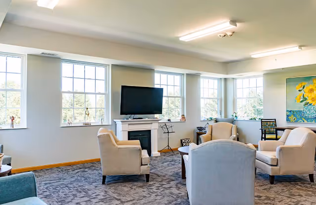 A bright living room with large windows letting in natural light. The room features a flat-screen TV mounted above a white fireplace, surrounded by beige upholstered armchairs arranged in a conversational grouping. There is a colorful painting on the wall and a music stand near the fireplace.