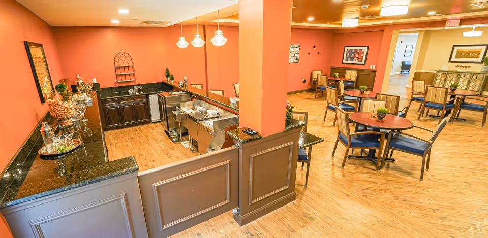 Interior view of a senior living facility dining area with several round tables and chairs arranged on a wooden floor. To the left, there is a serving counter with a black granite countertop, glassware, and decorative items. The walls are painted in warm tones of orange and beige, and pendant lights hang above the counter. The space appears clean, well-lit, and inviting.