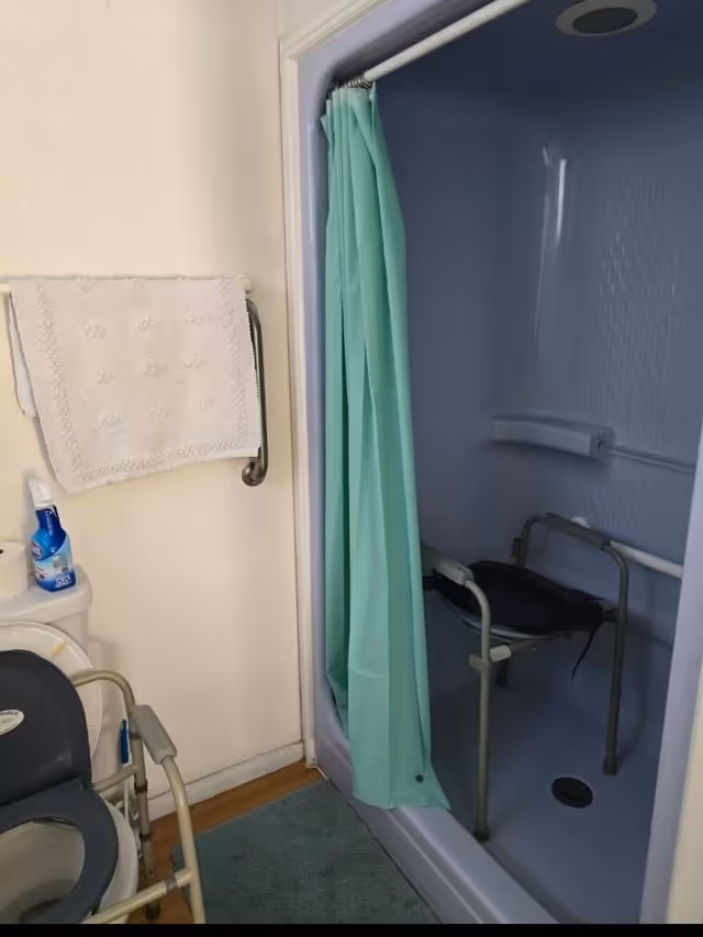 A bathroom with a shower area featuring a green shower curtain and a shower chair inside. There is a towel hanging on a rail above the toilet, which has a raised seat with armrests. A bottle of cleaning spray and a roll of toilet paper are placed on the toilet tank. The floor has a green bath mat.