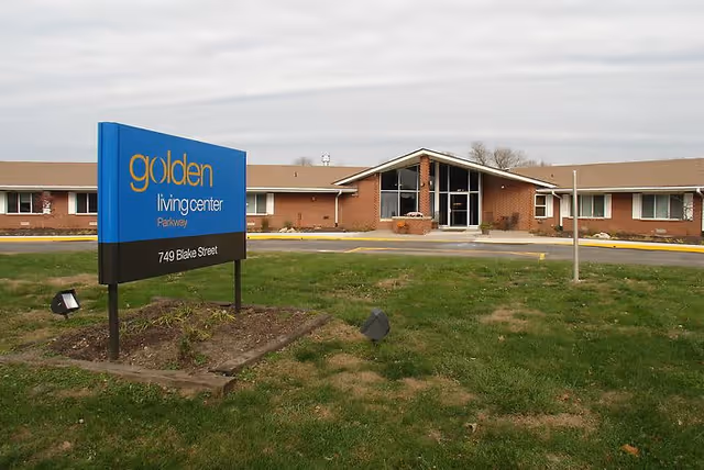 Exterior view of the Golden Living Center Parkway building with a blue and black sign in the foreground displaying the facility name and address 749 Blake Street. The building is a single-story brick structure with a peaked roof and large glass entrance.