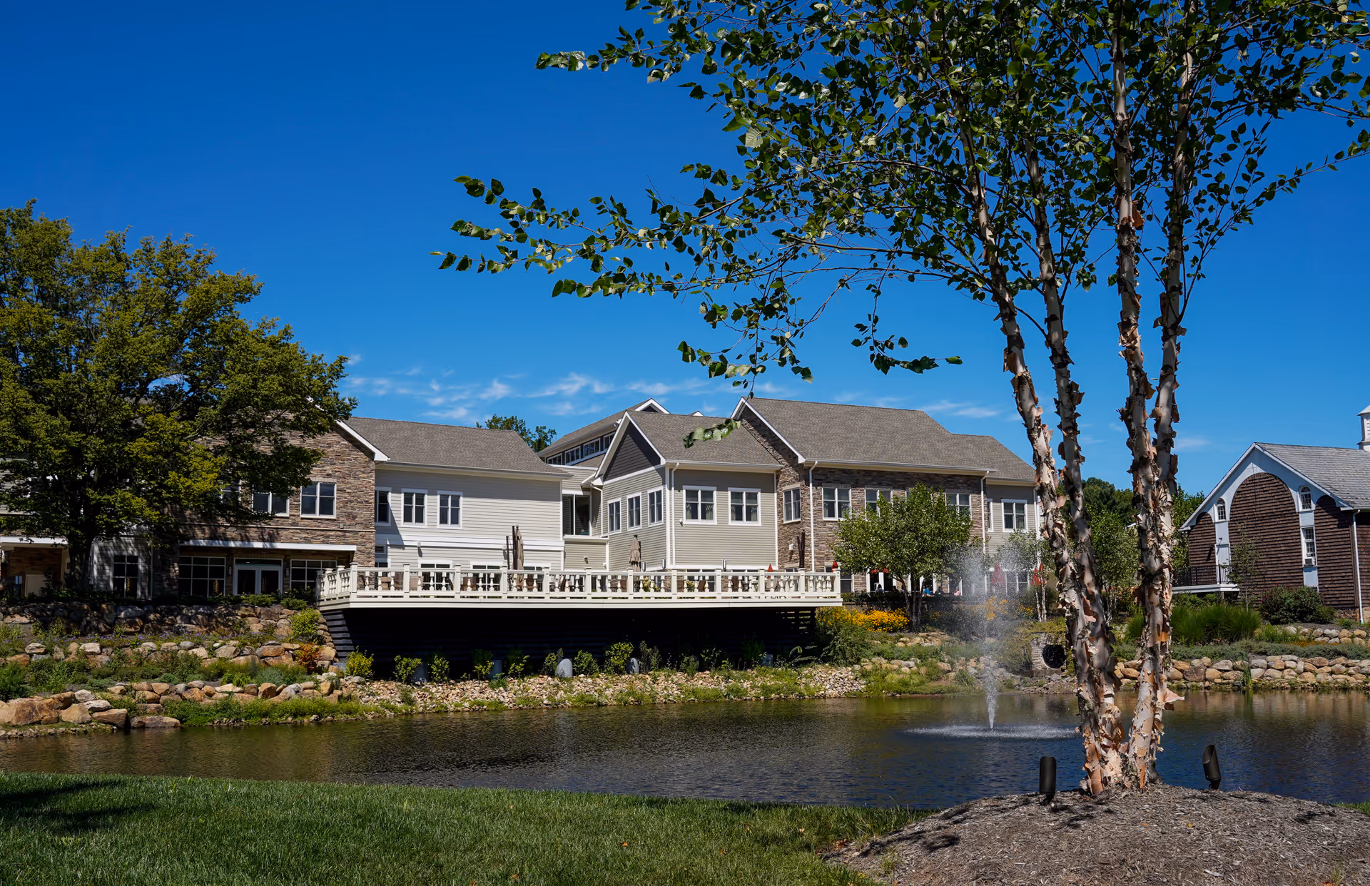 Exterior view of Pine Run Village senior living facility with a large pond and fountain in the foreground, surrounded by green grass and trees under a clear blue sky.