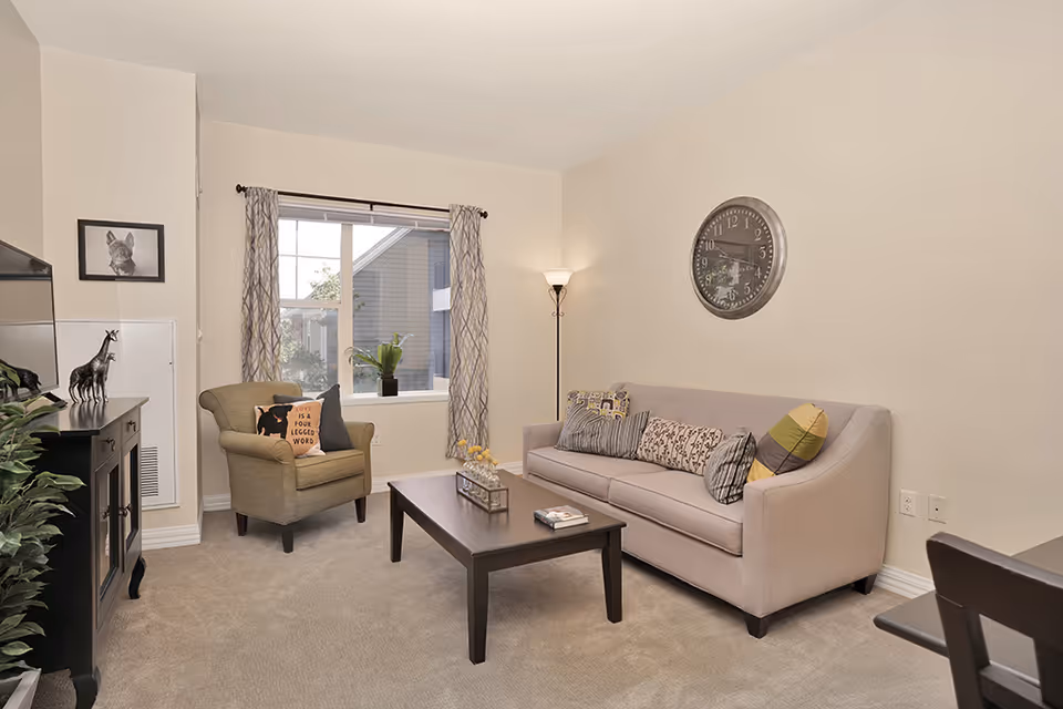 A cozy living room with a beige sofa adorned with multiple patterned pillows, a wooden coffee table with decorative items, an armchair with a cushion, a floor lamp, a wall clock, and a window with patterned curtains letting in natural light.