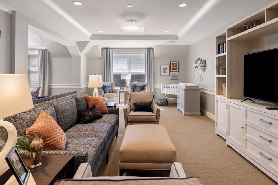 A cozy living room with a gray sofa adorned with orange and black pillows, a matching armchair, and a beige ottoman in the center. There are two armchairs with blue pillows near a window with gray curtains. A white dresser with a changing pad and a white crib are against the far wall. A large flat-screen TV is on a white entertainment unit with shelves and cabinets. The room is softly lit with table lamps and recessed ceiling lights.