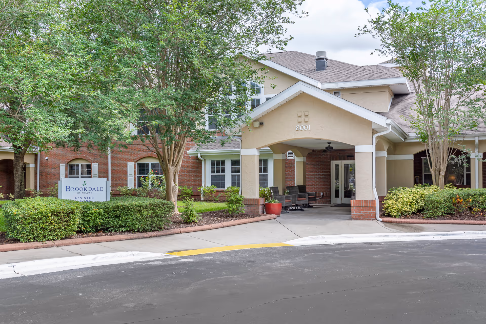 Exterior view of the entrance to Brookdale Dr. Phillips assisted living facility with a covered driveway, seating area, and surrounding greenery including trees and bushes.