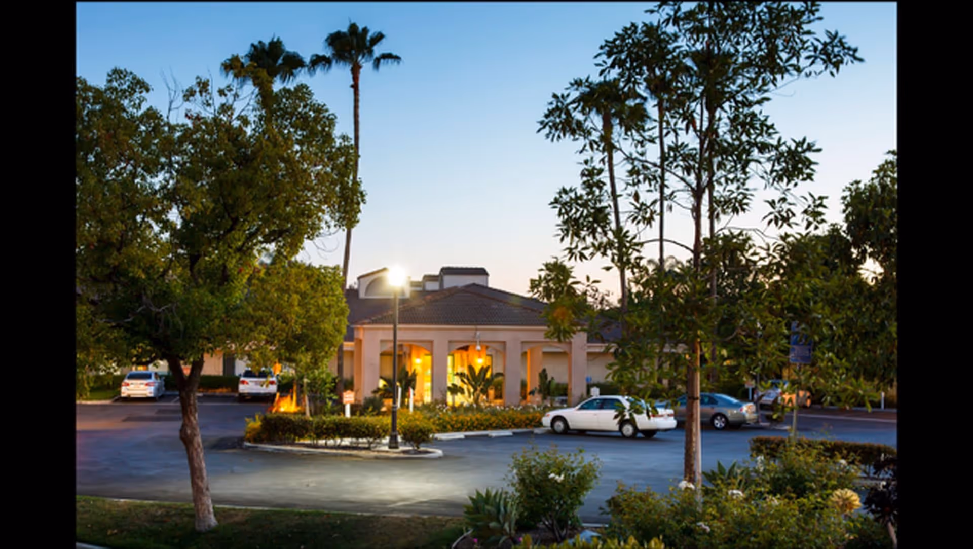 Exterior view of Park Vista Assisted Living and Memory Care facility at dusk, showing the building entrance with warm lighting, surrounded by trees, shrubs, and a parking lot with several cars.