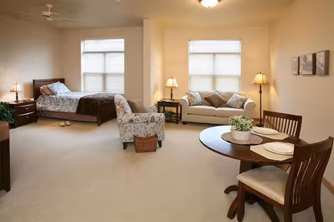 A spacious, well-lit room featuring a bed with bedside tables and lamps, a patterned armchair with a small basket, a white sofa with cushions, and a round dining table set with plates and chairs. The room has beige walls and carpet, with two large windows letting in natural light.