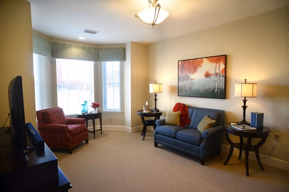 A cozy living room with a blue sofa adorned with two yellow patterned pillows and a red throw blanket. Two round wooden side tables with lamps flank the sofa. A large painting with autumn trees hangs above the sofa. To the left, a red patterned armchair sits near a bay window with blinds and a small round table holding decorative glass items and flowers. A TV is partially visible on the left side of the image.