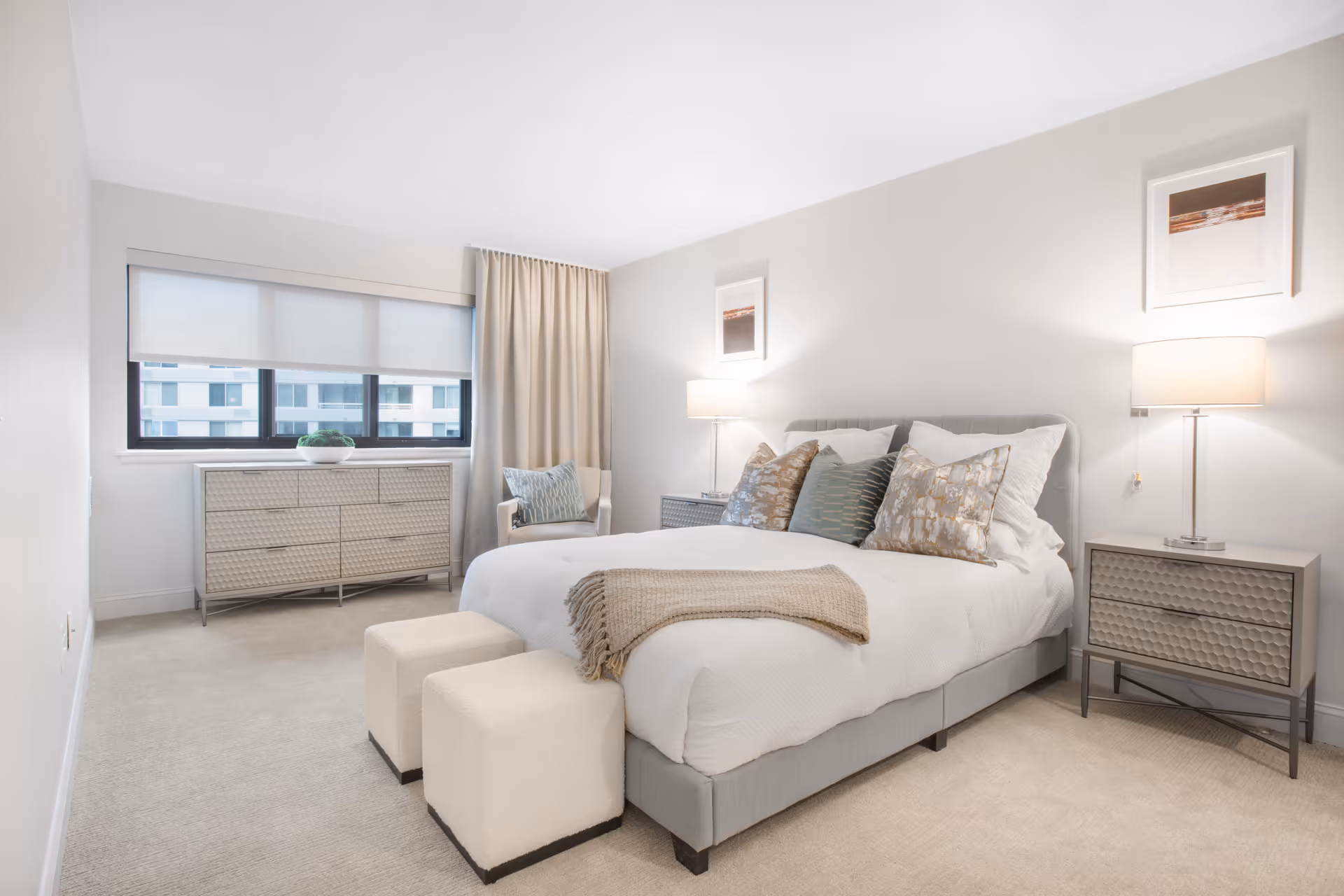 A bright, modern bedroom with a large bed featuring white bedding and multiple decorative pillows. Two beige ottomans are placed at the foot of the bed. On either side of the bed are matching nightstands with table lamps. A dresser with a plant on top is positioned under a window with a roller shade and beige curtains. The walls are light-colored and decorated with framed artwork.
