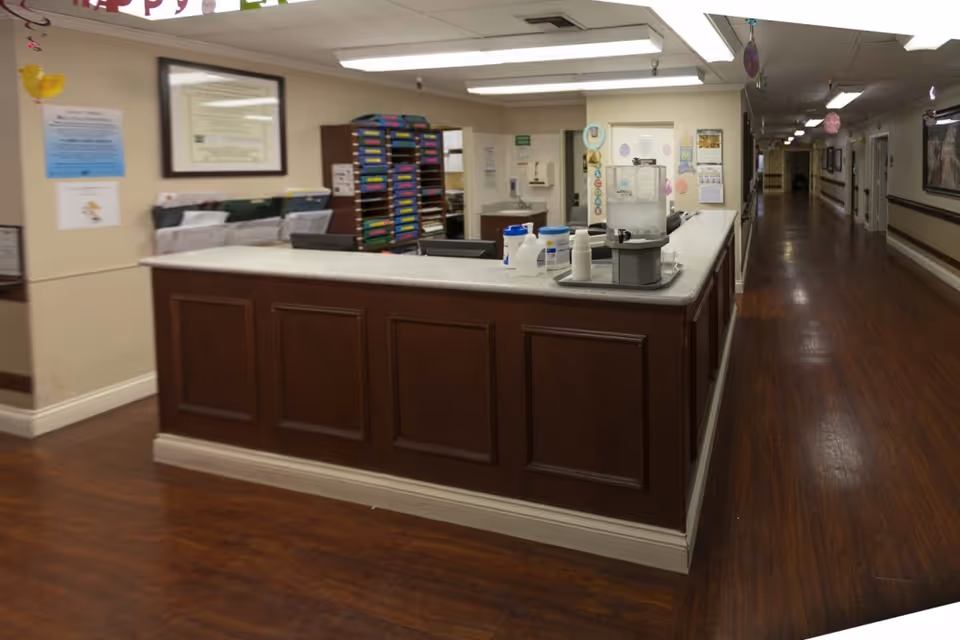 Reception desk area inside a healthcare facility with a long hallway extending to the right. The desk has a countertop with various containers and a water dispenser. Behind the desk are shelves with colorful folders and framed certificates on the wall. The floor is wooden and the walls are light-colored.