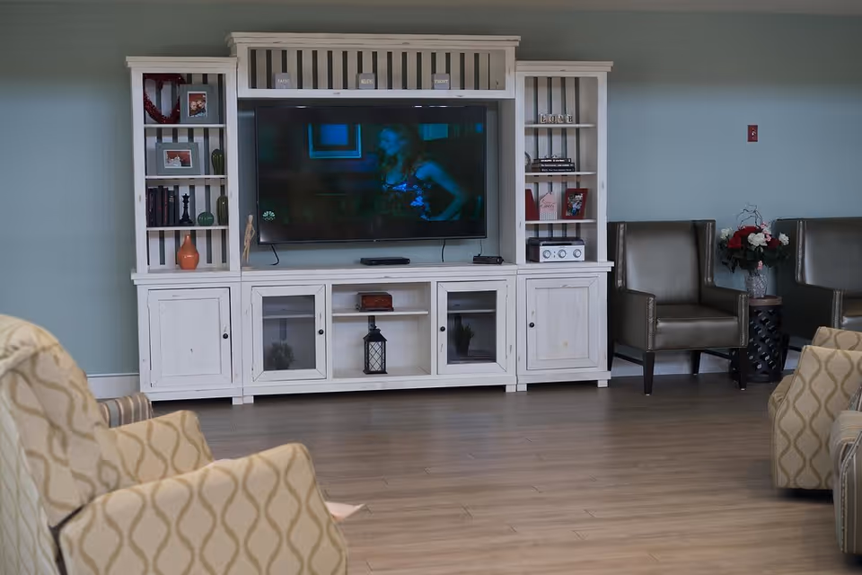 A living room area with a white entertainment center holding a flat-screen TV. The entertainment center has shelves with decorative items and books. There are patterned armchairs in the foreground and two brown leather chairs next to a small table with a flower arrangement on the right side. The floor is wooden and the walls are painted light blue.