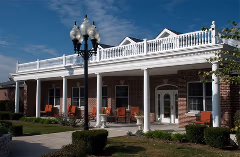 Exterior view of a brick building with a white porch railing and columns, several red cushioned chairs on the porch, a black vintage-style street lamp, and a clear blue sky with some clouds.