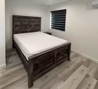 A simple bedroom with a wooden bed frame and a mattress, a small nightstand next to the bed, a window with a black and white striped blind, and light-colored wooden flooring.