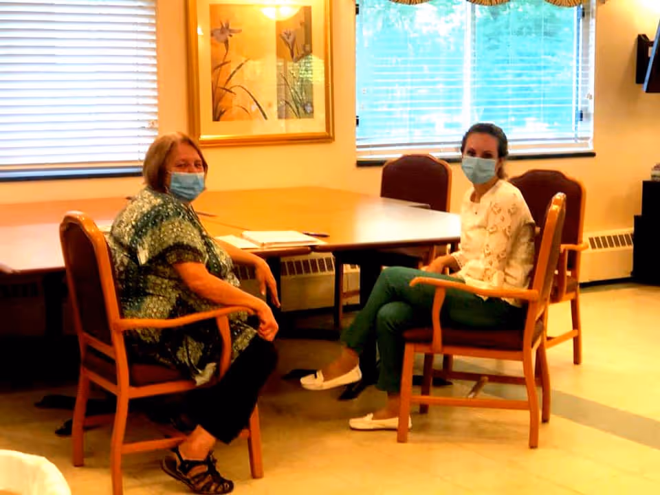 Two women wearing face masks sitting on wooden chairs around a large wooden table in a well-lit room with windows covered by blinds and a framed floral picture on the wall.