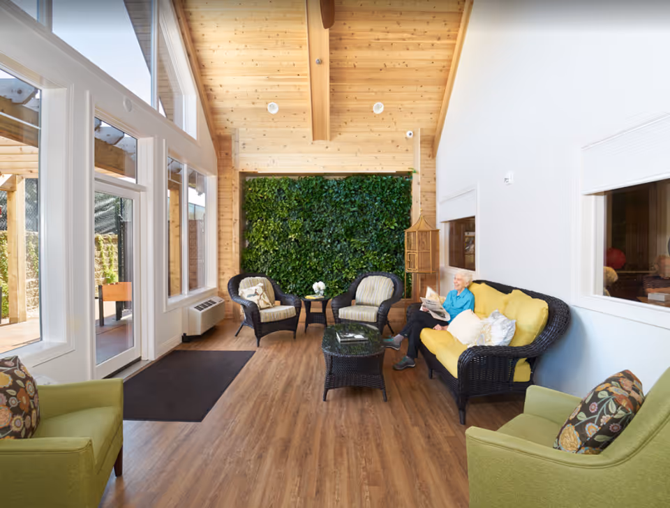 A bright and cozy sitting area with wooden floors and a wooden ceiling. The room features large windows and glass doors letting in natural light. There are two green armchairs with patterned cushions, two wicker chairs with striped cushions, and a wicker sofa with yellow cushions. A small black coffee table and side table hold decorative items. A green vertical garden is mounted on the far wall. An elderly woman is sitting on the sofa reading a newspaper.