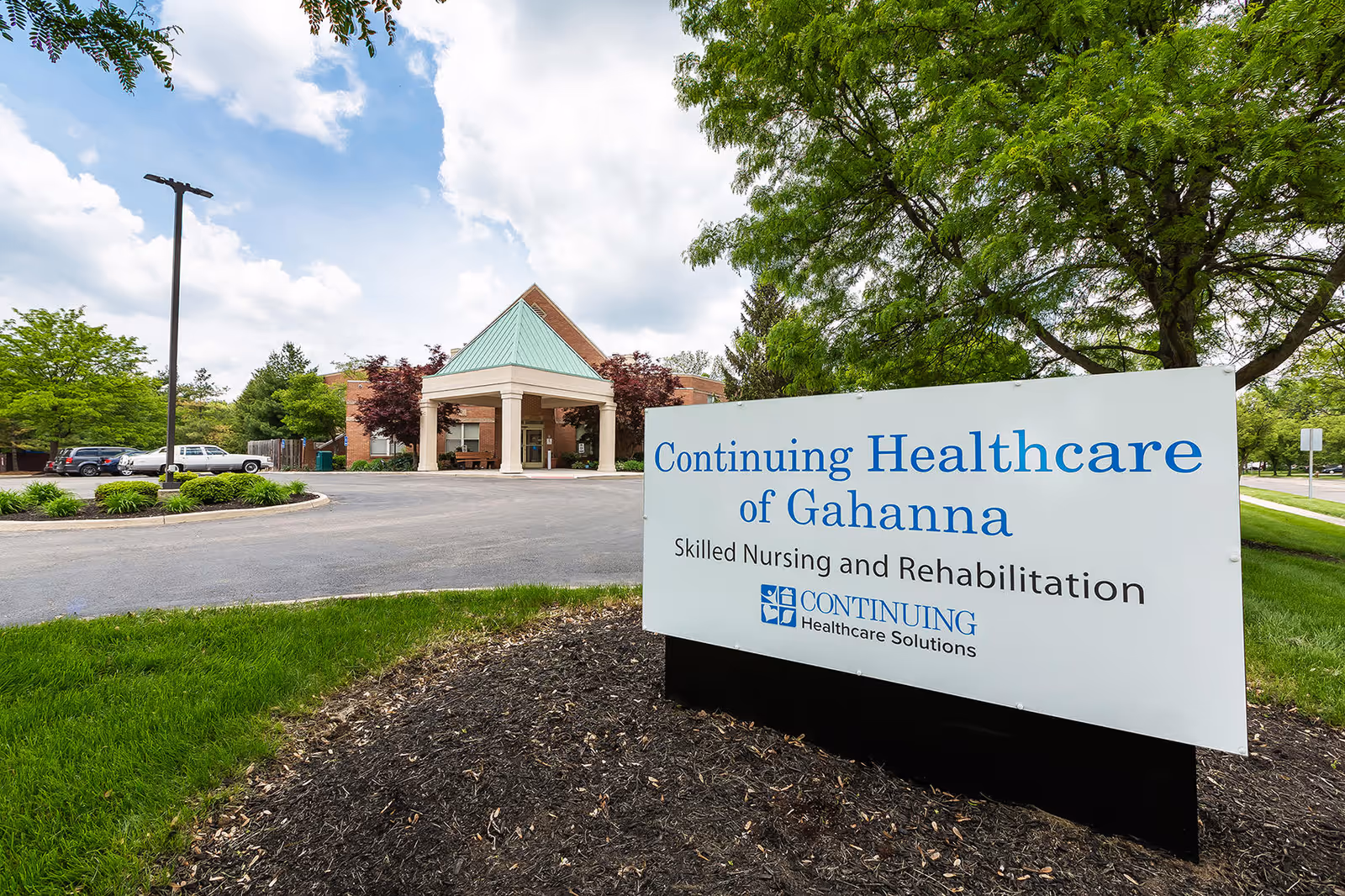 Exterior view of Continuing Healthcare of Gahanna building with a driveway, parking area, and a large sign in the foreground that reads 'Continuing Healthcare of Gahanna Skilled Nursing and Rehabilitation'. The building has a covered entrance with a green roof and is surrounded by trees and greenery under a partly cloudy sky.
