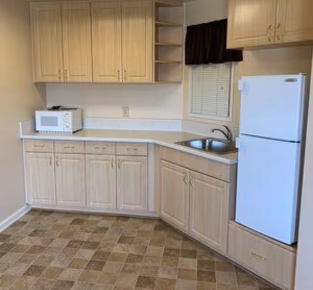 A small kitchen area with light wood cabinets, a white countertop, a white microwave on the counter, a stainless steel sink under a window with a dark valance, and a white refrigerator. The floor has a checkered pattern with brown and beige tiles.
