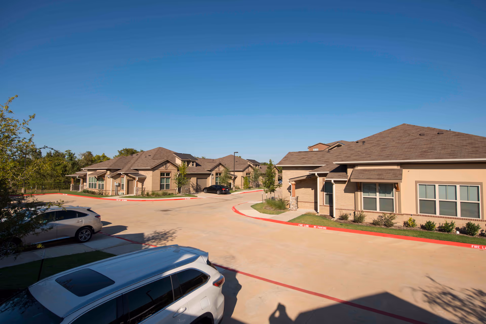 Exterior view of a senior living facility with multiple single-story buildings, a clear blue sky, and several parked cars along the driveway.