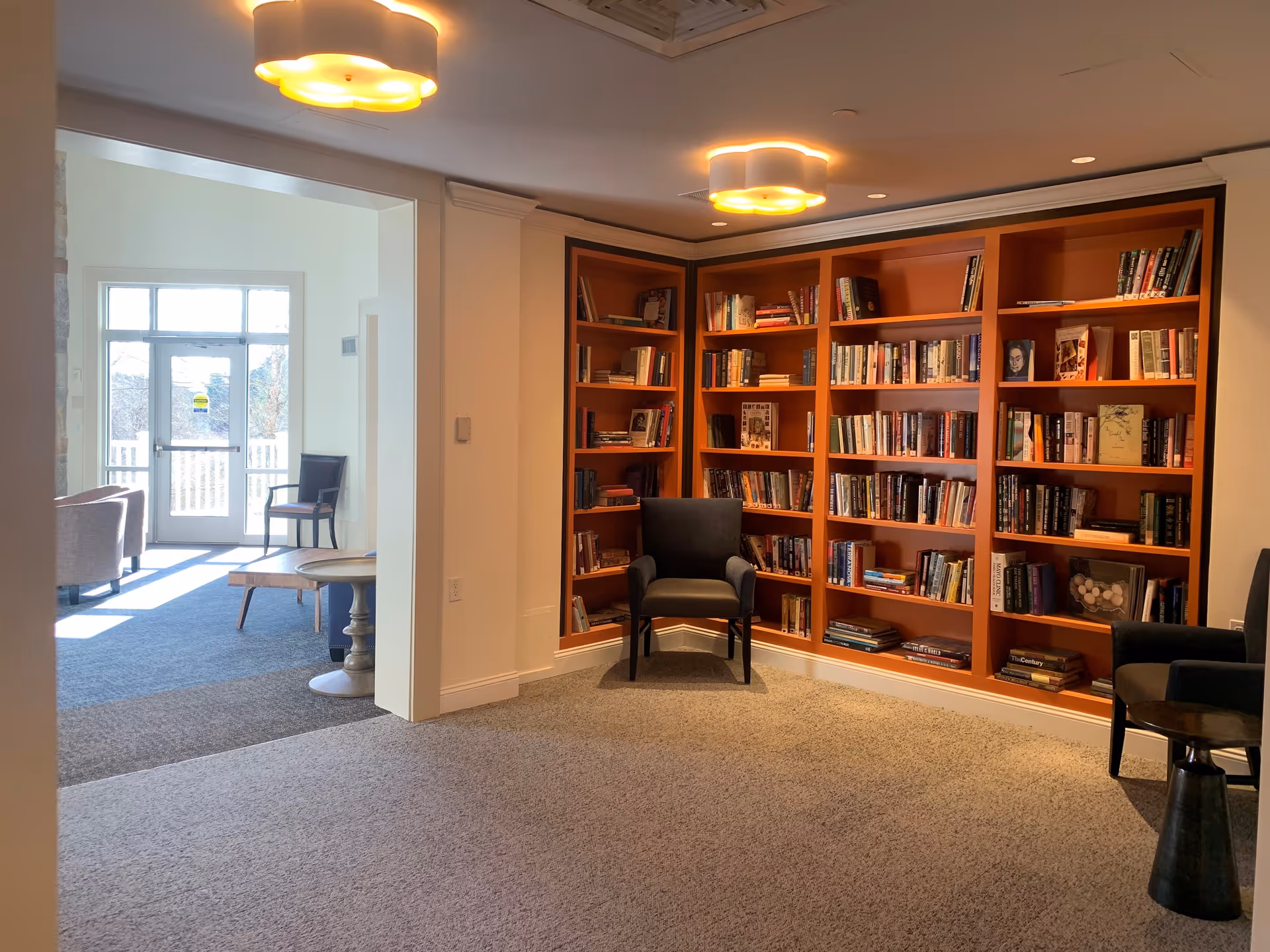 A cozy reading nook with built-in orange bookshelves filled with books, two black armchairs, and a small round side table. The area is carpeted and well-lit with ceiling lights. Adjacent to the nook is a bright room with large windows and a glass door leading outside, furnished with additional chairs and tables.