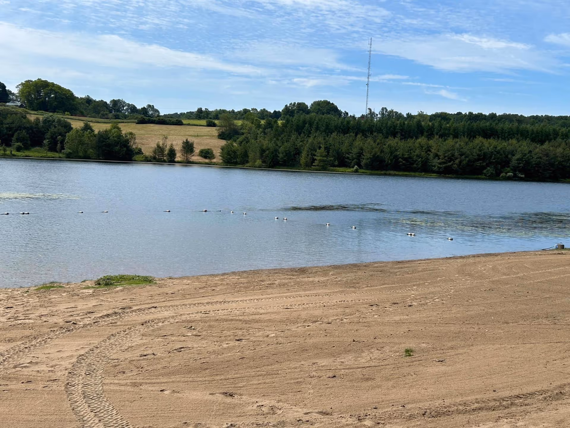 A sandy beach area with tire tracks in the foreground leading to a calm lake. Across the lake, there are green trees and grassy hills under a partly cloudy blue sky.