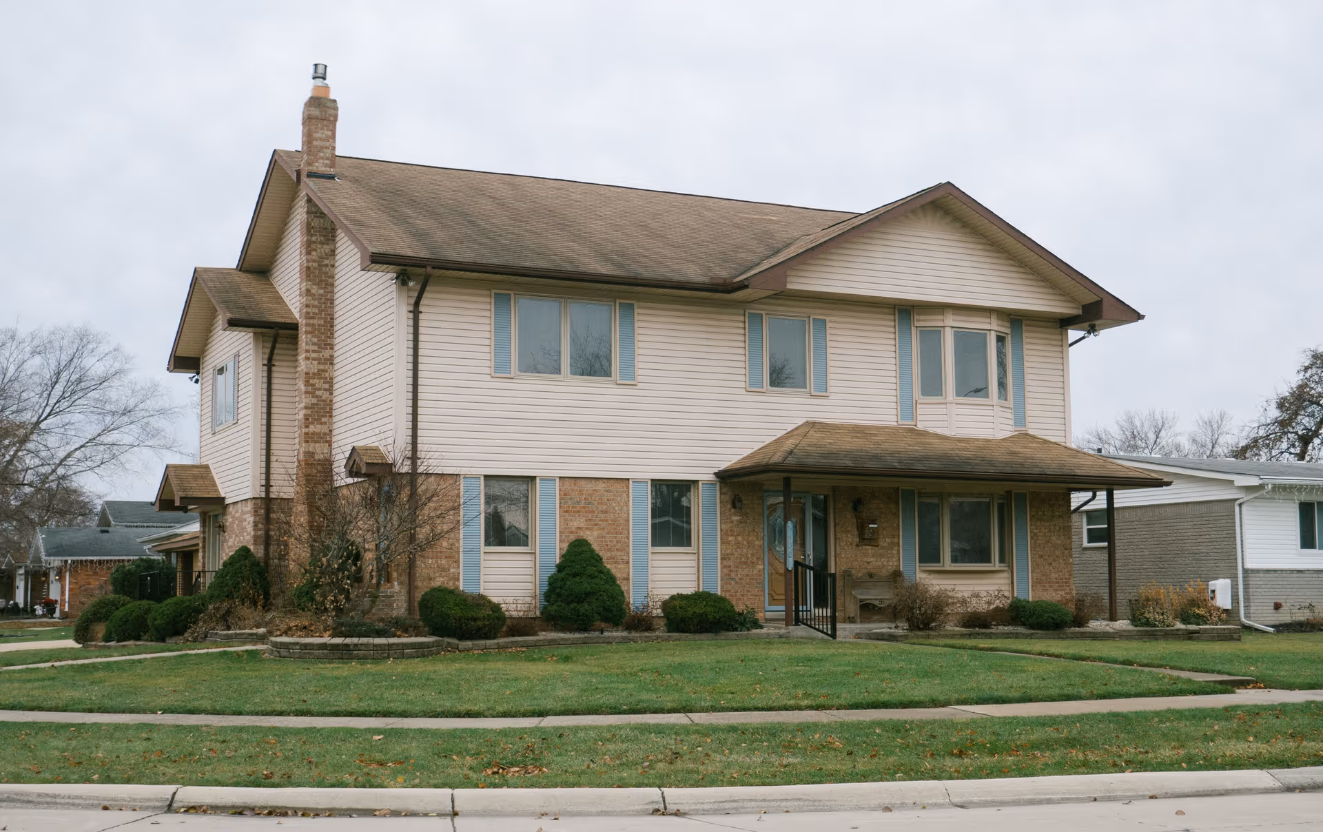 Exterior view of a two-story residential building with beige siding and brick accents. The building has multiple windows with blue shutters, a covered front porch with a wooden door, and a chimney on the left side. The lawn is green with trimmed bushes and a sidewalk in front.