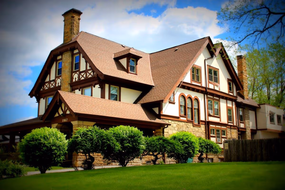 Exterior view of a large, multi-story building with Tudor-style architecture, featuring brown wooden beams, white walls, and a steeply pitched roof. The building is surrounded by green bushes and a well-maintained lawn under a partly cloudy blue sky.