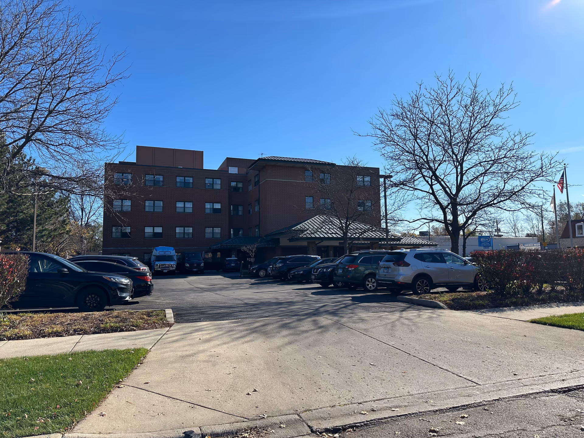 Exterior view of a four-story brick building with a green metal roof entrance, surrounded by parked cars and leafless trees under a clear blue sky.