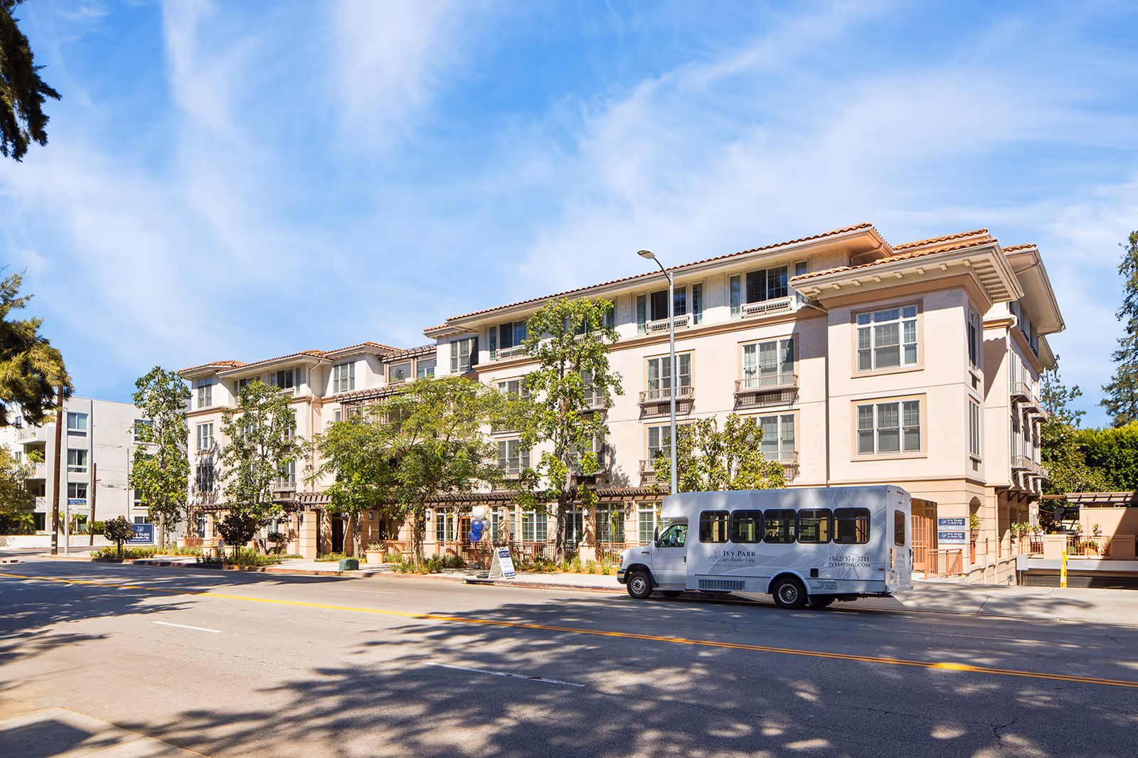 Exterior view of a multi-story senior living facility building named Ivy Park at Studio City, with a shuttle bus parked in front on a sunny day with a blue sky and some trees around.