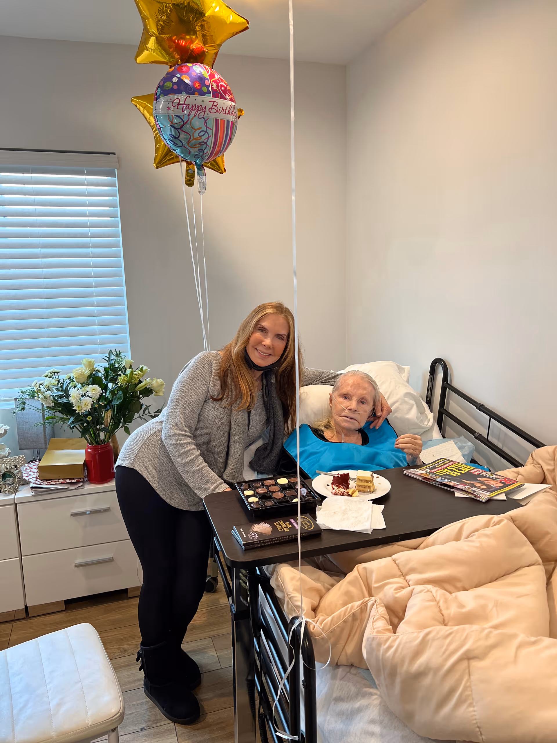 An elderly woman lying in a hospital bed with a blue bib, accompanied by a younger woman standing beside her. The bed has a tray table with a plate of cake, chocolates, and magazines. There are birthday balloons and a bouquet of flowers on a nearby cabinet in a well-lit room.