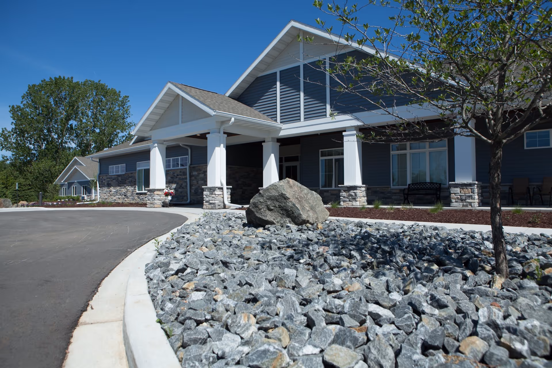 Exterior view of a single-story building with blue siding and stone accents, featuring a covered entrance supported by white columns. In the foreground, there is a landscaped area with large rocks and a tree, and a curved driveway leading to the entrance under a clear blue sky.