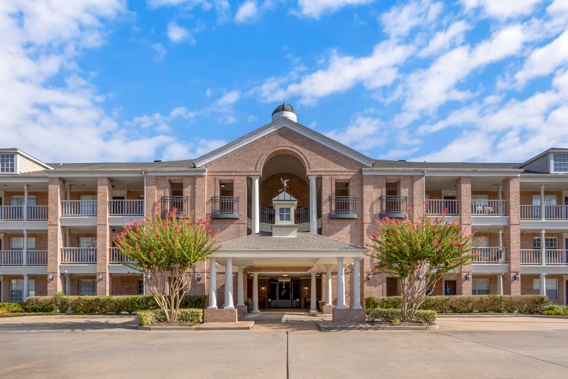Front exterior view of a three-story brick building with white columns and balconies, featuring a covered entrance with a small cupola on top. Two flowering trees flank the entrance, and the sky is partly cloudy.