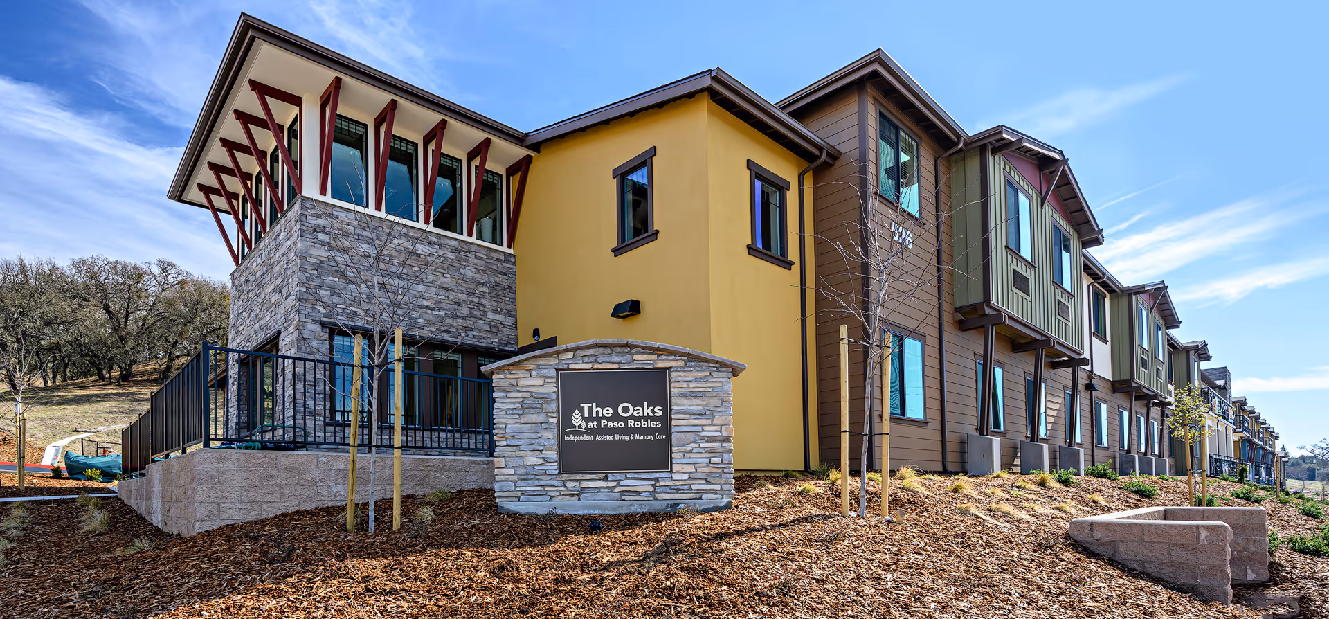 Exterior view of The Oaks at Paso Robles senior living facility showing a modern multi-story building with stone and yellow siding, a sign with the facility name, young trees, and a clear blue sky.