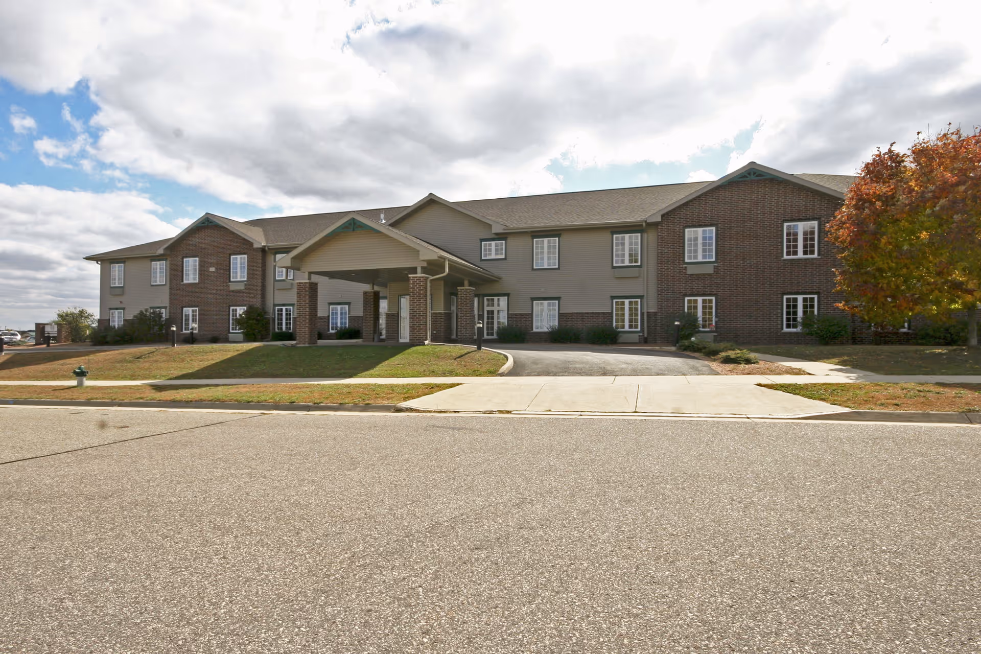 Front exterior view of a two-story senior living apartment building with a covered entrance, brick and siding facade, several windows, a tree with autumn foliage on the right, and a paved road in the foreground under a partly cloudy sky.