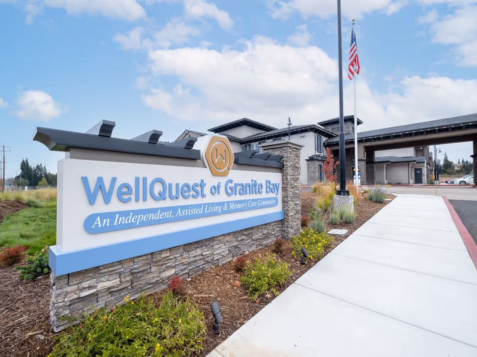 Entrance sign for WellQuest of Granite Bay, an independent, assisted living and memory care community, with the facility building and an American flag in the background under a partly cloudy sky.