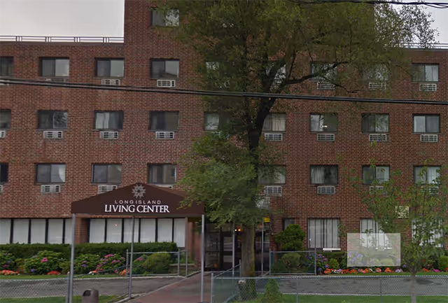 Exterior view of a multi-story brick building with multiple windows, some with air conditioning units. The entrance has a brown awning with the text 'Long Island Living Center'. There are trees, bushes, and flowers in front of the building, along with a fenced area and a sidewalk leading to the entrance.
