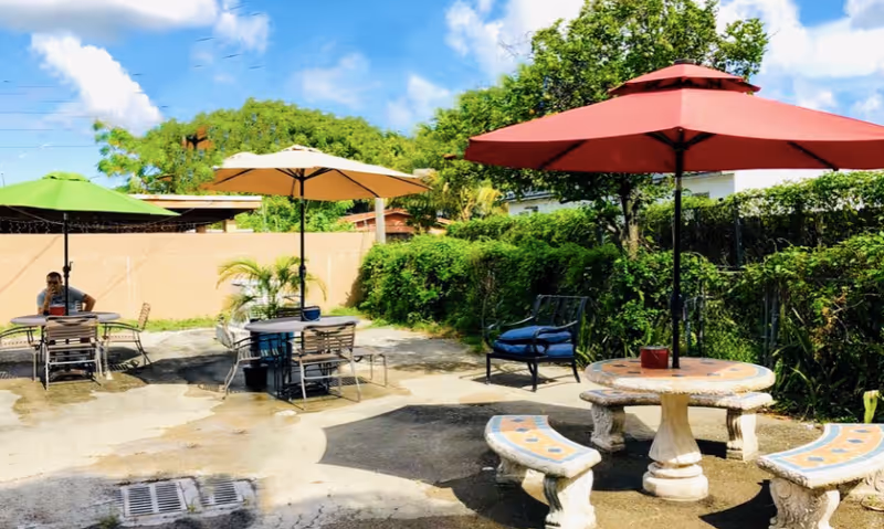 Outdoor patio area with several tables and chairs under colorful umbrellas, surrounded by greenery and a clear blue sky. One person is sitting at a table under a green umbrella.