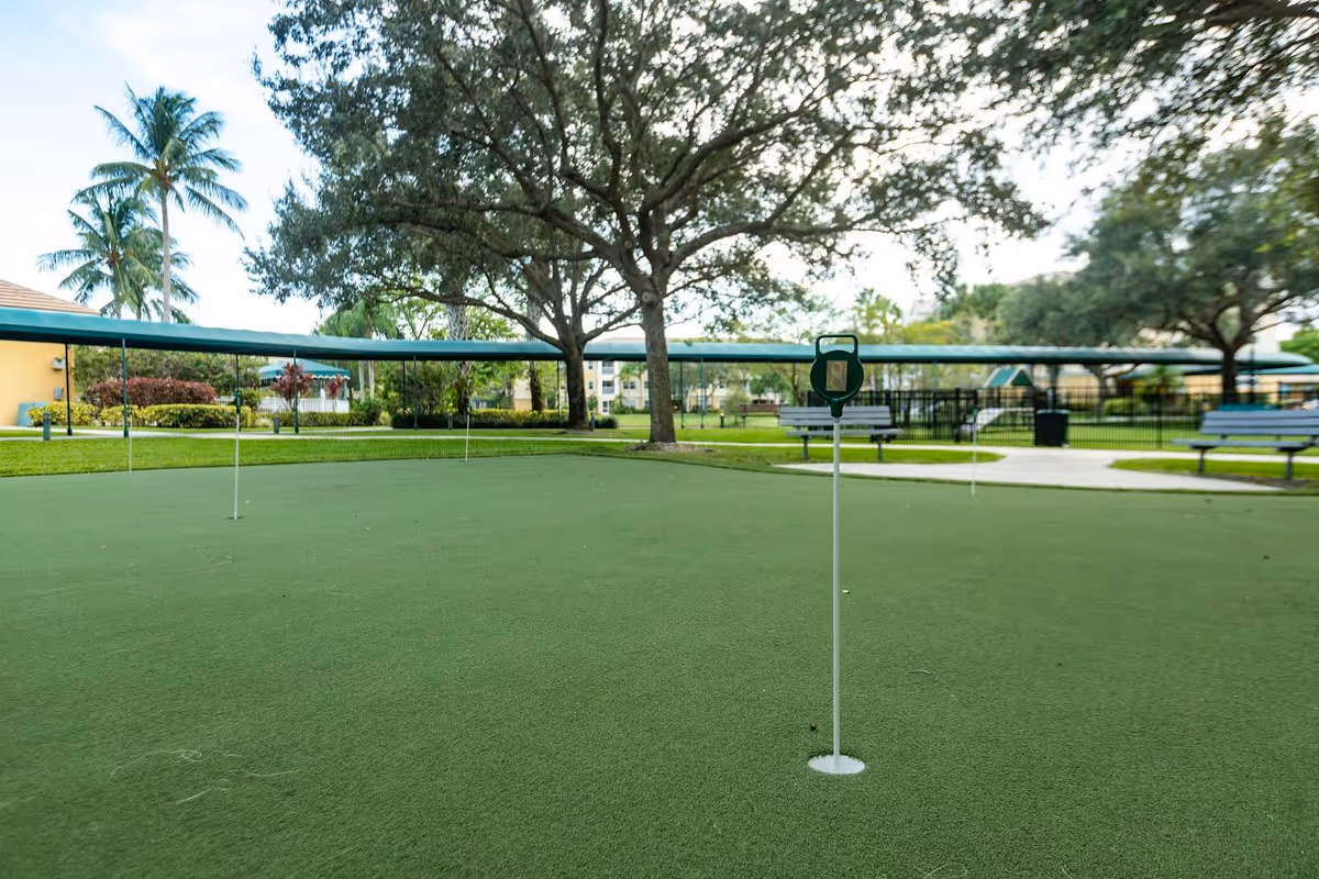 Outdoor putting green with several golf holes and flags, surrounded by trees, benches, and covered walkways in a senior living community.