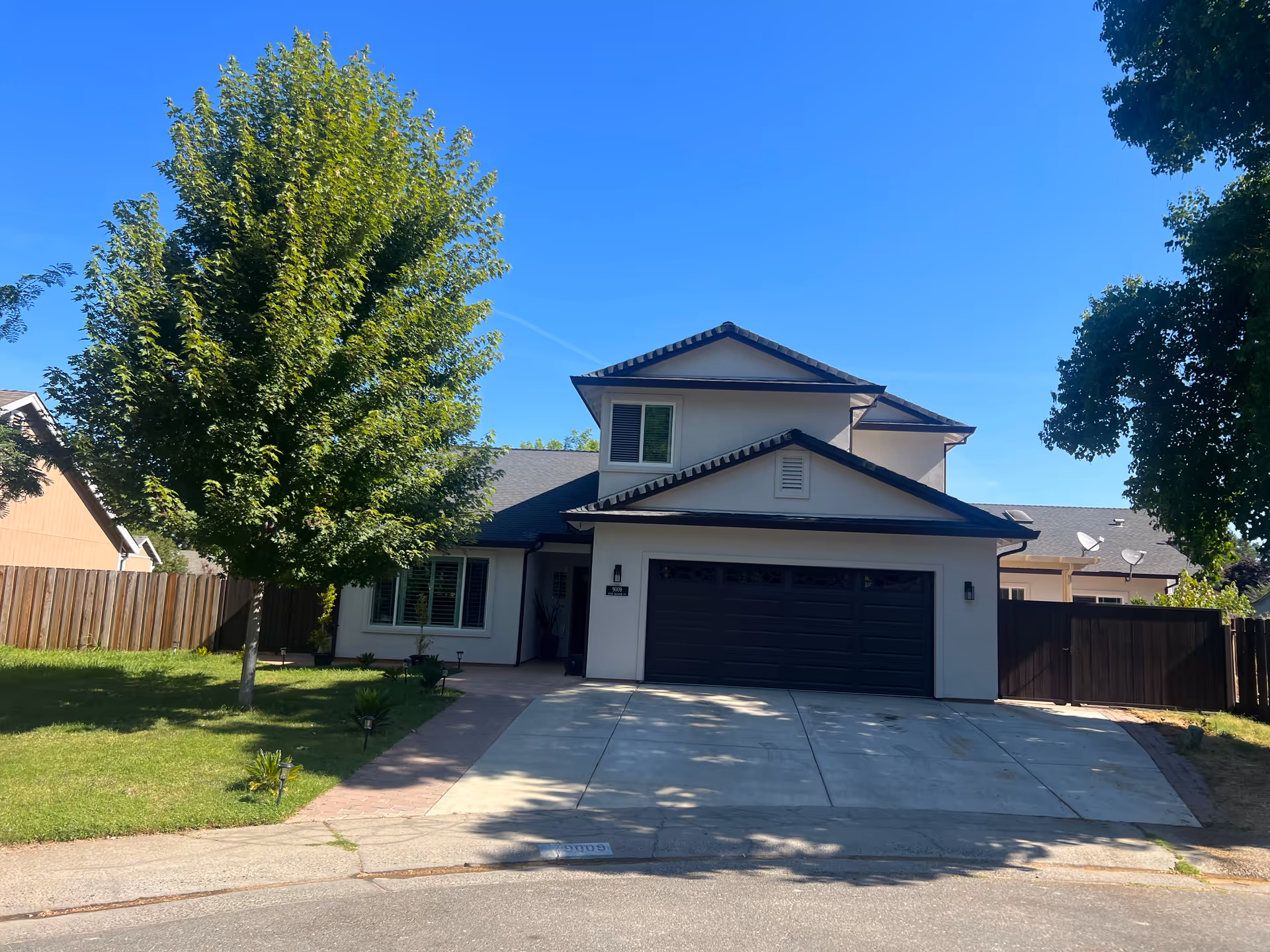 Front exterior view of a two-story residential building with a dark garage door, white walls, and a sloped roof. There is a large green tree on the left side of the driveway and a wooden fence surrounding the property. The sky is clear and blue.