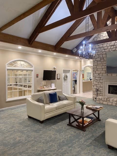 A spacious living room area in a senior living facility with a high vaulted ceiling featuring exposed wooden beams and a chandelier. The room includes a white sofa with blue and gray pillows, a wooden coffee table with books and a vase of flowers, a large stone fireplace with a mounted TV above it, and a wall with arched windows and a door leading to another room.