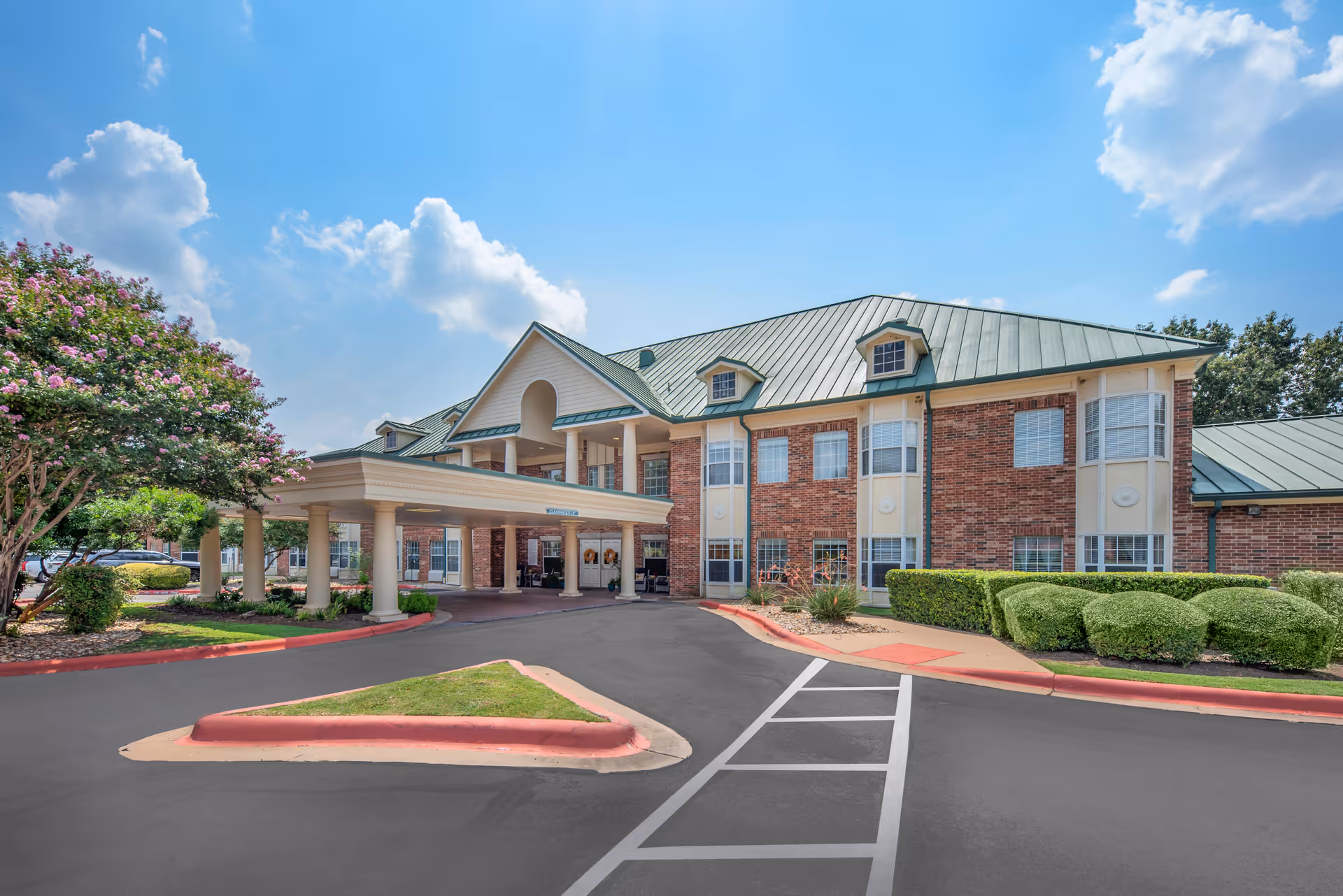 Exterior view of Brookdale Beckett Meadows senior living facility showing a two-story brick building with a green metal roof, a covered entrance with columns, neatly trimmed bushes, a tree with pink flowers, and a clear blue sky with some clouds.