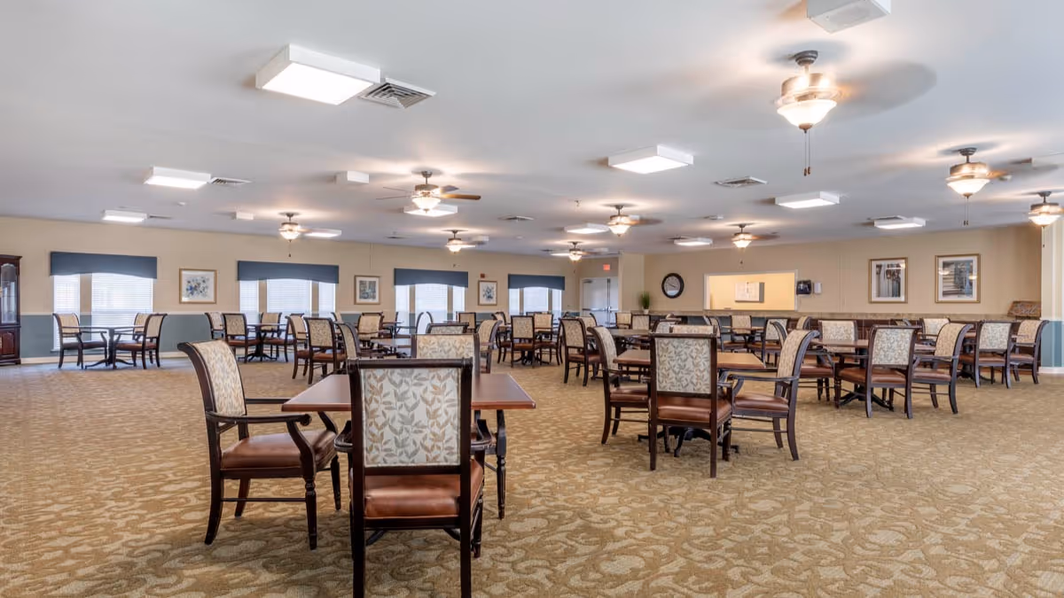 A spacious dining room with multiple wooden tables and cushioned chairs arranged neatly. The room has beige walls, patterned carpet flooring, several ceiling fans with lights, and windows with blue valances allowing natural light to enter. There are framed pictures on the walls and a clock near a service counter area.