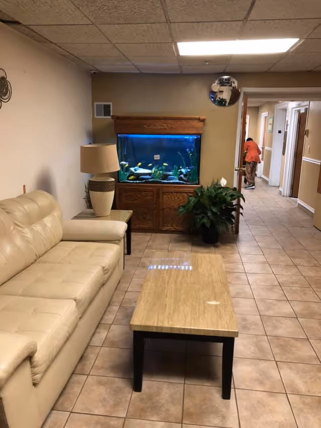 A cozy indoor seating area with a beige leather couch, a wooden coffee table, a side table with a lamp, and a large fish tank set in a wooden cabinet against the wall. There is a green potted plant next to the fish tank. The floor is tiled, and a hallway is visible in the background with a person walking away.