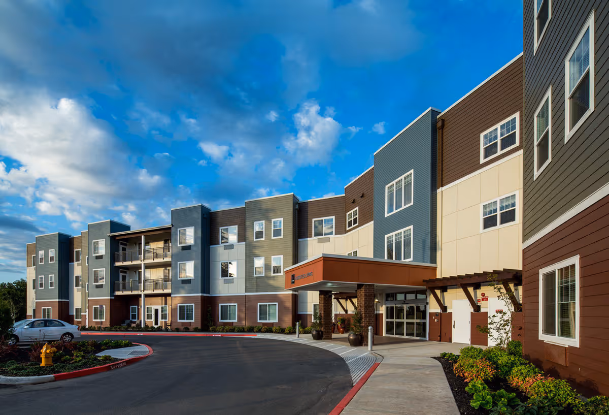 Exterior view of a modern three-story assisted living facility building with multiple windows, a covered entrance, landscaped greenery, and a clear blue sky with clouds.