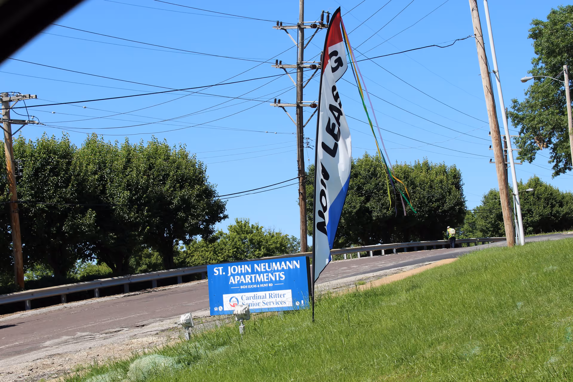 Outdoor view of a grassy area with a blue sign for St. John Neumann Apartments and a tall flag that says 'Now Leasing' against a clear blue sky with trees and utility poles in the background.