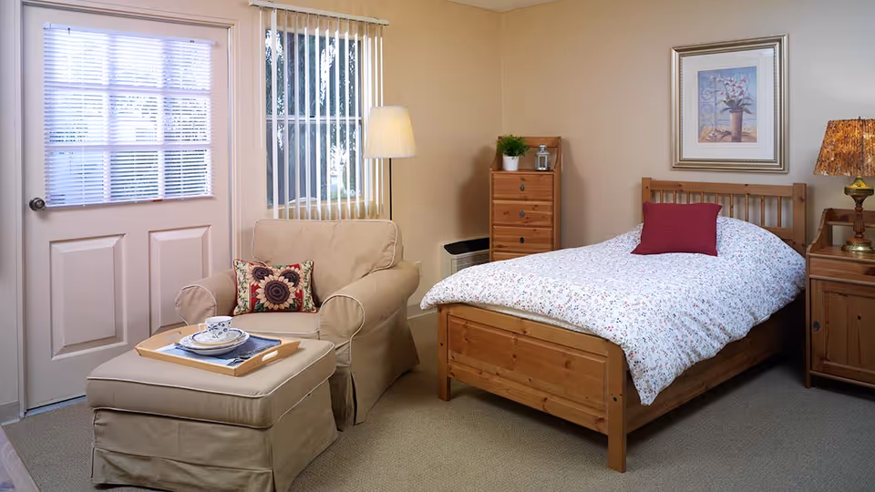 A cozy bedroom with a wooden bed featuring a floral bedspread and a red pillow. Next to the bed is a wooden nightstand with a lamp. There is a wooden dresser with a plant and a lantern on top. A beige armchair with a floral cushion and matching ottoman is placed near a door and window with blinds. The walls are painted light beige and a framed floral artwork hangs above the bed.