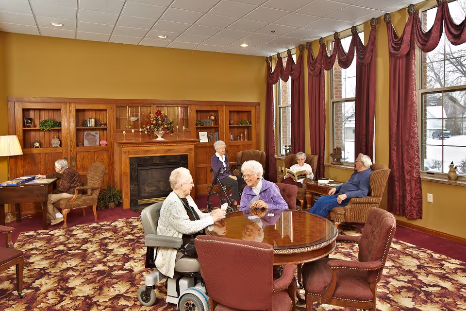 A cozy common area in a senior living facility with five elderly people engaged in various activities. Two women are sitting at a round wooden table playing cards, one woman is sitting at a desk reading, and two others are seated in armchairs near large windows with red curtains, one reading a book and the other relaxing. The room features a fireplace with wooden built-in shelves, a patterned carpet, and warm yellow walls.