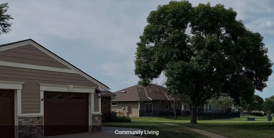 Exterior view of a residential community area with a garage, a large tree, green lawns, and multiple single-story buildings under a partly cloudy sky.