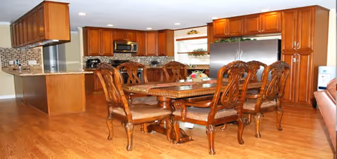 A spacious kitchen and dining area featuring wooden cabinetry, a large stainless steel refrigerator, a microwave, and a tiled backsplash. In the center is a wooden dining table with six ornate chairs. The floor is hardwood, and there is a window with blinds above the kitchen counter.