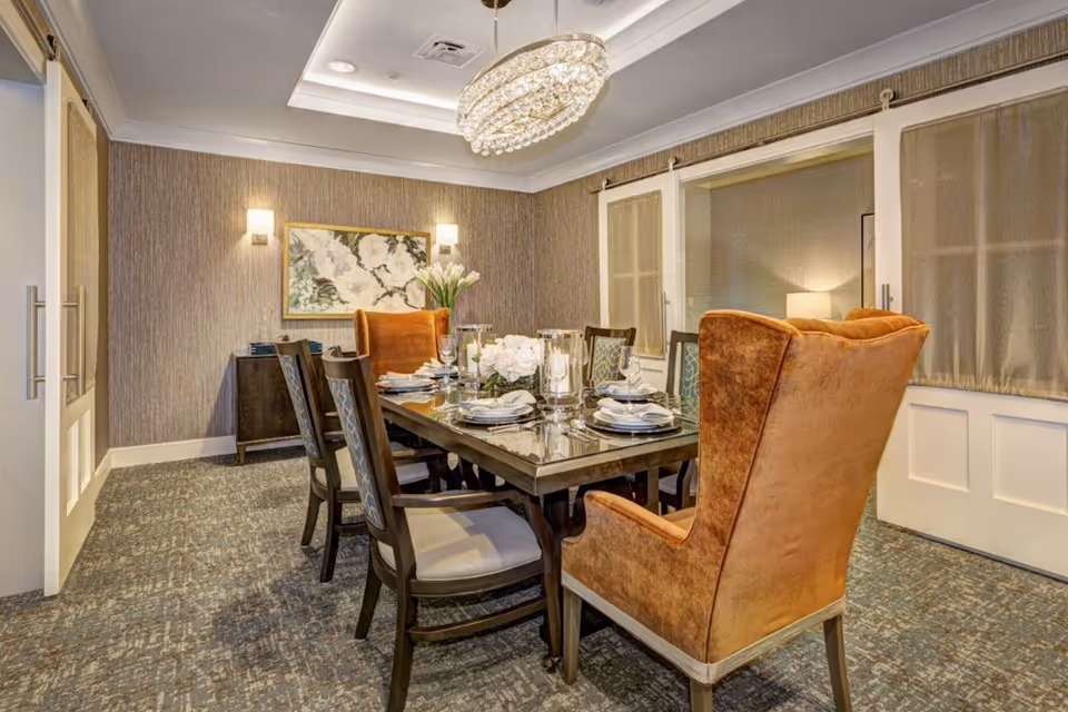 A well-lit dining room with a rectangular glass-top table set for six people. The table is surrounded by six chairs, including two large, plush, orange wingback chairs at the ends. The room features textured wallpaper, a patterned carpet, a floral painting on the wall, and a modern chandelier hanging from the ceiling. There are sliding doors with curtains on one side of the room.